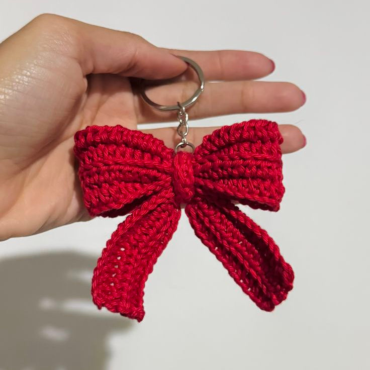 Red crocheted bow keychain held in a hand against a plain background